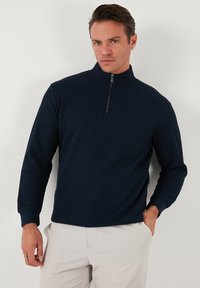 Navy ribbed long-sleeve pullover with a high collar and half zip. Features a structured texture and fitted cuffs. Light-colored pants.