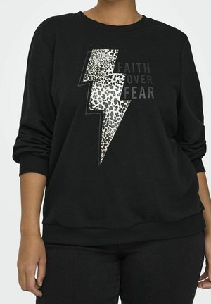 Personne portant un sweat-shirt noir avec un éclair imprimé léopard et le texte « La foi plutôt que la peur » sur le devant, manches retroussées.