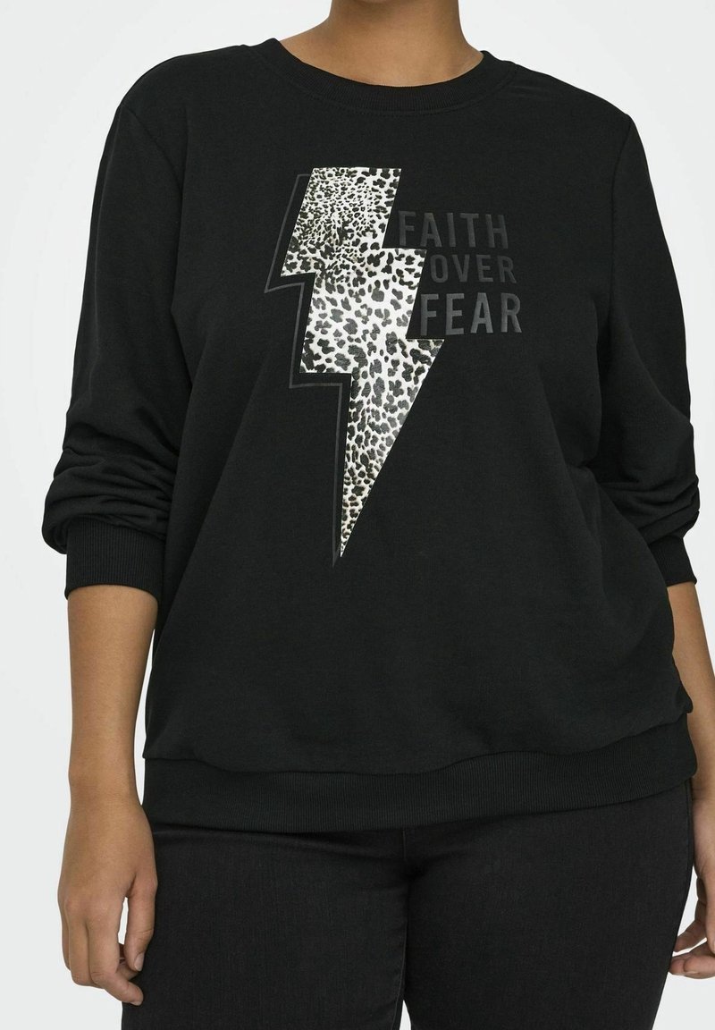 Personne portant un sweat-shirt noir avec un éclair imprimé léopard et le texte « La foi plutôt que la peur » sur le devant, manches retroussées.