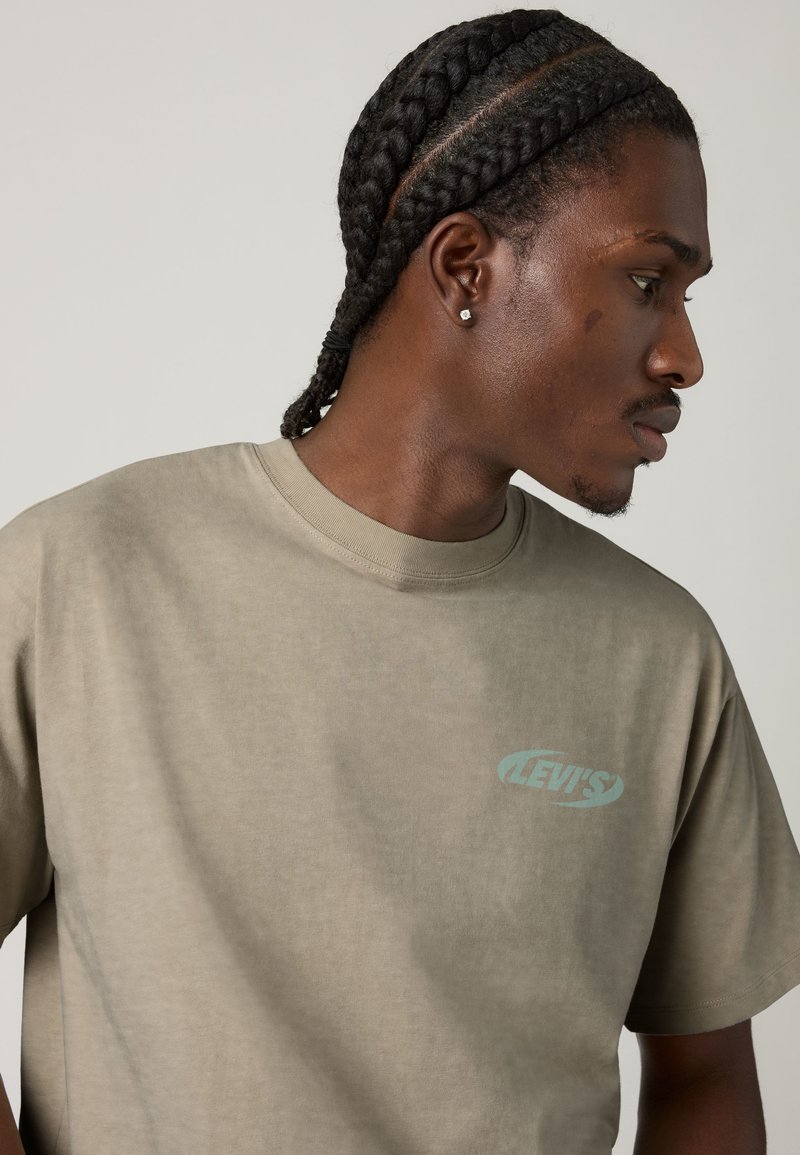 Homme avec des tresses cornrow et une boucle d'oreille en diamant, portant un t-shirt Levi's vert discret, regardant vers sa droite sur un fond uni.