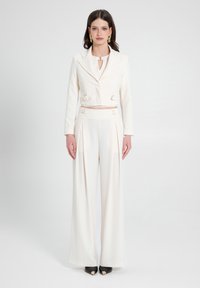 Blazer bianco cropped con fibbie laterali, abbinato a pantaloni a gamba larga. Tessuto liscio con una vestibilità su misura e una leggera lucentezza. Scarpe nere a punta.