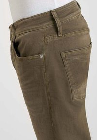 Pantalon en denim marron exposé sur un mannequin, montrant des poches latérales et arrière avec des coutures visibles et des passants de ceinture.