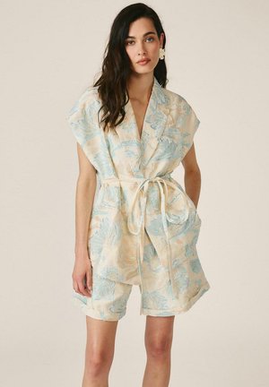 Mujer con cabello largo y oscuro que lleva una chaqueta sin mangas beige y azul con estampado floral y cinturón, y pantalones cortos a juego, de pie con la mano en el bolsillo.