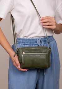 Olivově zelená kožená crossbody taška s kapsou na zip na přední straně, hladkým povrchem, štíhlým designem a nastavitelným popruhem.