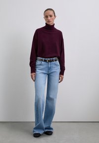 Woman standing wearing a dark purple turtleneck sweater, light blue flared jeans with a black belt, and black shoes against a plain background.