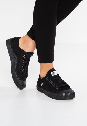 Personne portant des baskets en toile noires basses avec des lacets noirs et une étiquette de marque blanche sur la languette, associées à un pantalon noir ajusté.