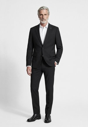 Älterer Mann mit grauem Haar und Bart, der einen schwarzen Anzug, weißes Hemd und schwarze Schuhe trägt und vor einem einfarbigen hellen Hintergrund steht.