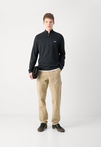 Sudadera negra con cremallera y un pequeño logo, pantalones cargo beige y zapatillas negras. El modelo sostiene un bolso de mano negro. Fondo liso.