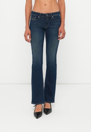 Kobieta nosząca ciemnoniebieskie jeansy bootcut i czarne czółenka w szpic, stojąca na jednolitym białym tle.