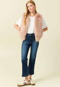 Jasnoróżowa kamizelka z futerka, biała koszulka, niebieskie dżinsy z wysokim stanem i beżowe sneakersy z bordowymi akcentami, na neutralnym tle.