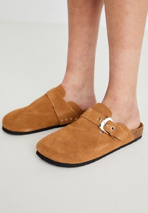 Brown suede slip-on clogs with a wide strap, silver buckle detail, and flat black rubber soles, showcasing a smooth texture and rounded shape.