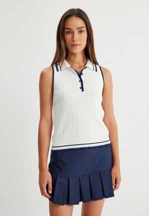 Polo sin mangas de punto en color blanco con detalles en azul marino y cinco botones. Combinado con una falda corta plisada en azul marino. Textura suave; diseño ajustado.