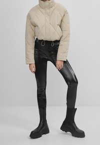 Beige quilted cropped jakke med høj krave, kombineret med tætsiddende sorte glansfulde leggings og kraftige sorte ankelstøvler.