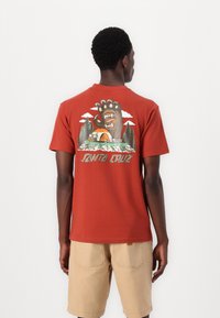 Red short-sleeve t-shirt featuring a graphic of a bear paw and a camping scene, with green trees and clouds. Plain back. Cotton material.