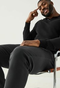 Juodas polo marškinėliai ilgomis rankovėmis ir tamsios kelnės, sėdintys ant chromuoto kėdės su rudos odos elementais, išryškinantys lygius paviršius ir prigludusią konstrukciją.