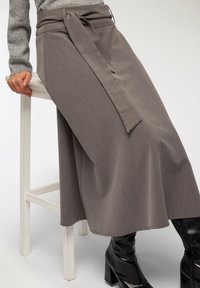 Midi spódnica z teksturowanej szarej tkaniny z wiązaniem w talii, w połączeniu z dopasowanymi czarnymi kozakami do kolan, oparta na białym stołku.