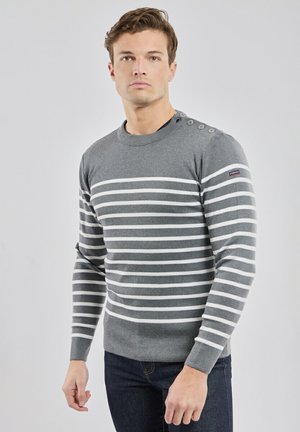 Mand iført en grå sweater med hvide horisontale striber og knapdetaljer på skulderen, kombineret med mørkeblå jeans.