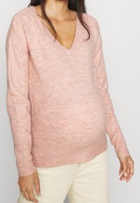 Pull rose clair à col en V avec un tricot doux et texturé. Il présente des manches longues et une coupe décontractée, conçu pour s'adapter à un ventre qui s'arrondit.