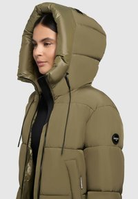 Olive green padded jacket with oversized hood, zip closure, and elastic drawstrings. Features a logo patch on the shoulder and side pockets.