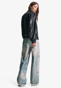Schwarze Kunstleder-Bomberjacke mit gerippten Bündchen, kombiniert mit hellblauen Denim-Hosen, die eine ausgewaschene Optik und mehrfarbige abstrakte Muster aufweisen.