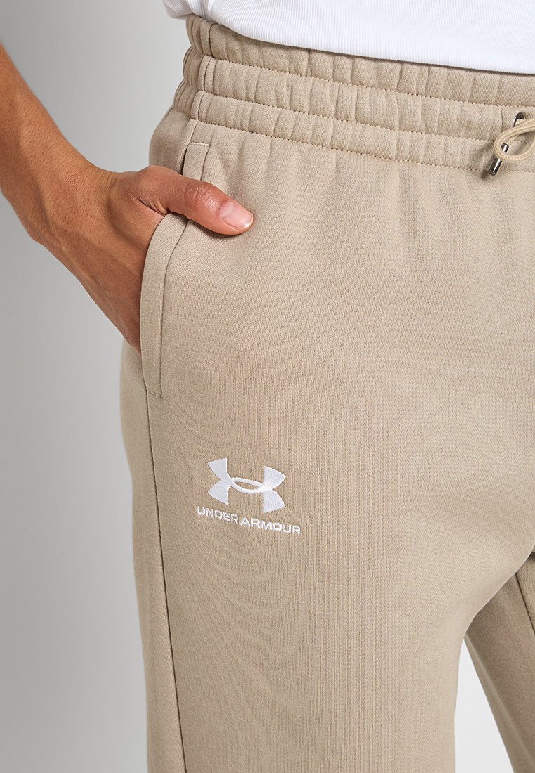 Zbliżenie na osobę w beżowych spodniach dresowych Under Armour z elastycznym pasem i boczną kieszenią, ręka spoczywająca w kieszeni.