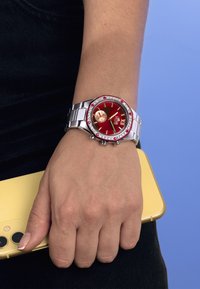Reloj de acero inoxidable plateado con un dial rojo redondo, bisel acentuado con cristales y funciones de cronógrafo, llevado en una mano que sostiene un teléfono amarillo.