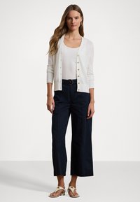 Balta jakas ar zelta pogām, vilkta virs baltas topa ar korseti, apvienota ar tumšām platajām biksēm un gaišām sandalēm ar dekoratīviem akcentiem.