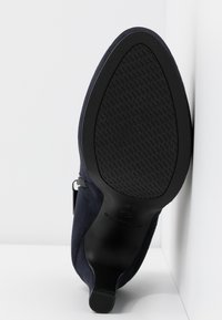 Semelle d'une chaussure noire à talon pointu avec grip en caoutchouc, reposant verticalement contre un mur d'angle blanc et le sol.