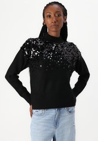 Maglione nero con un collo alto e spalle adornate da paillettes nere e argentate, abbinato a jeans azzurri.