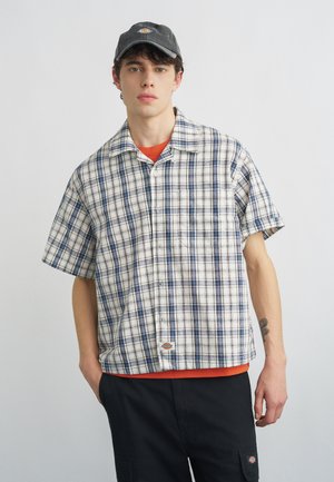 Młody mężczyzna w czarnej czapce, białej i niebieskiej koszuli w kratę z krótkim rękawem, pomarańczowej podkoszulce i czarnych spodniach z bocznymi kieszeniami.