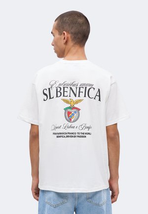 Homme aux cheveux courts portant un t-shirt blanc SL Benfica avec texte et emblème dans le dos, associé à un jean bleu clair.
