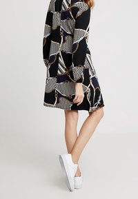 Svart klänning med ett geometriskt mönster av kedjor och ränder i vitt och guld, med långa ärmar och lös passform, ihop med vita sneakers.