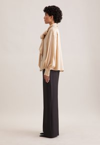 Blusa beige con maniche a sbuffo e polsini con bottoni, abbinata a pantaloni neri a vita alta e décolleté verdi appuntiti. Design semplice e elegante.