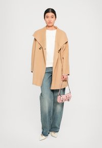 Moteris stovi vilkėdama didelį kupranugario spalvos paltą, baltą sweatshirt, plačiąsias kelnes, baltus smailianosius batus, laikydama mažą rožinę rankinę šalia savęs.