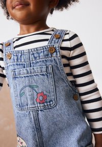 Denim-overalls med en frontlomme med blomsterdesign, træknapper og en langærmet trøje med sorte og hvide striber.