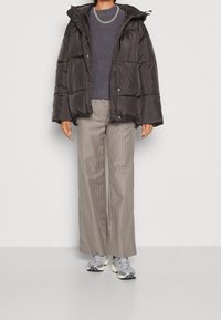 Chaqueta de plumas negra con capucha, diseño acolchado, combinada con un suéter gris texturizado y pantalones beige a rayas; zapatillas grises completan el look.
