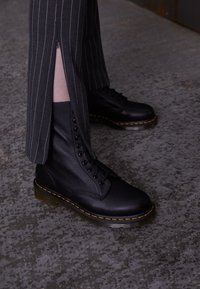 Botas de cuero negro con cordones y tobillo alto, con ojales, costuras amarillas y suela translúcida. Llevadas con pantalones a rayas.