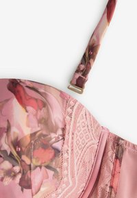 Rožinė gėlių liemenėlė su nėrinių detalėmis ir reguliuojama petnešėlė ant baltos fono, rodanti puodelio ir šoninio pulto artimą vaizdą.