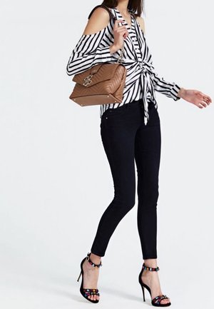 Blouse rayée décolletée avec un nœud à l'avant, jean skinny noir, et talons hauts à brides chevilles ornés de perles multicolores, portant un sac à main marron.