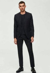 Μαύρο κοστούμι με ταιριαστό σακάκι και στενές παντελόνες από λεία ύφασμα. Συνδυασμένο με μαύρη μπλούζα με στρογγυλή λαιμόκοψη και μαύρα παπούτσια.