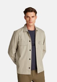 Giacca beige chiaro dal design elegante, con chiusura a bottone, due tasche frontali e un logo discreto sul petto. Tessuto leggero.