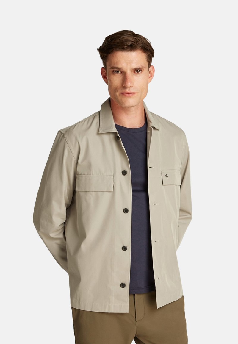 Giacca beige chiaro dal design elegante, con chiusura a bottone, due tasche frontali e un logo discreto sul petto. Tessuto leggero.