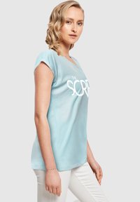 Lyseblå bomulds T-shirt med korte ærmer, der har et stort hvidt grafisk tryk med teksten "THE SCRIPT." Præsenteret med hvide bukser.