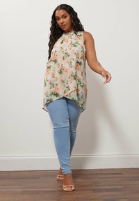 Blusa senza maniche floreale di colore crema con motivi arancioni e verdi, orlo asimmetrico. Abbinata a jeans attillati blu chiaro e sandali nude.