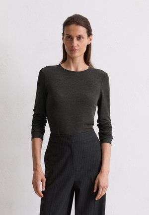 Ženska z dolgimi rjavimi lasmi, oblečena v prilegajočo temnosivo dolgotrajno majico z dolgimi rokavi in visokimi pinstripe temnimi hlačami, stoji ob preprosti beli steni.