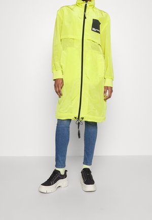 Persoon die een lange neongele jas met zwarte rits draagt, blauwe spijkerbroek, limoengroene sokken en zwarte sneakers met plateauzool, tegen een effen achtergrond.