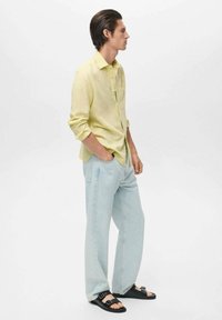 Camisa de manga larga color amarillo claro, pantalones de mezclilla azul claro de corte relajado y sandalias negras con tiras. Diseño sencillo, tonos neutros y detalles mínimos.