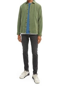 Camisa de pana verde sobre una camiseta azul, combinada con vaqueros negros y zapatillas deportivas de color claro. Cuenta con cierre de botones y cuello.