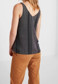 Donna che indossa un top senza maniche nero con strisce verticali bianche a pois e pantaloni marroni, vista di spalle contro un muro bianco semplice.