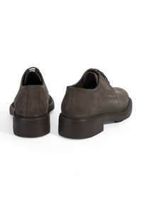 Une paire de chaussures habillées pour hommes en daim marron foncé avec des semelles noires épaisses, montrées de l'arrière sur un fond blanc.